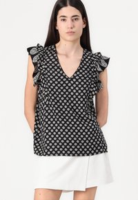 Top nero senza maniche con scollatura a V, motivo floreale bianco e dettagli arricciati sulle spalle, abbinato a una gonna bianca.