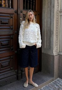 Pull en tricot blanc avec un motif floral, associé à un short en jean foncé et des ballerines beige, devant un fond de porte en bois.