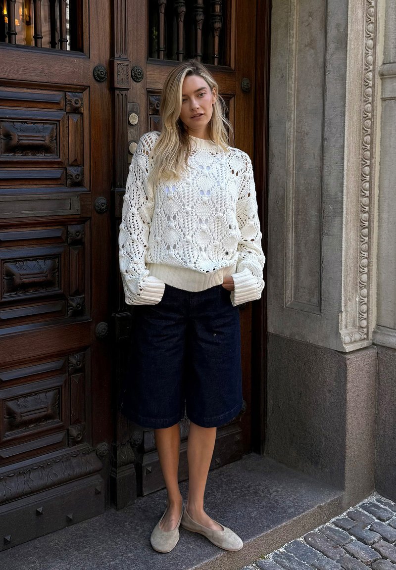 Pull en tricot blanc avec un motif floral, associé à un short en jean foncé et des ballerines beige, devant un fond de porte en bois.