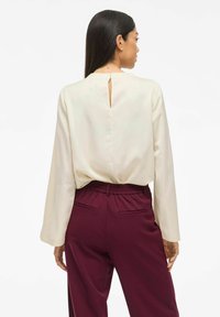 Blusa lunga a maniche con foro a chiave sul retro, di colore crema, abbinata a pantaloni borgogna con elastico in vita e tasche laterali.