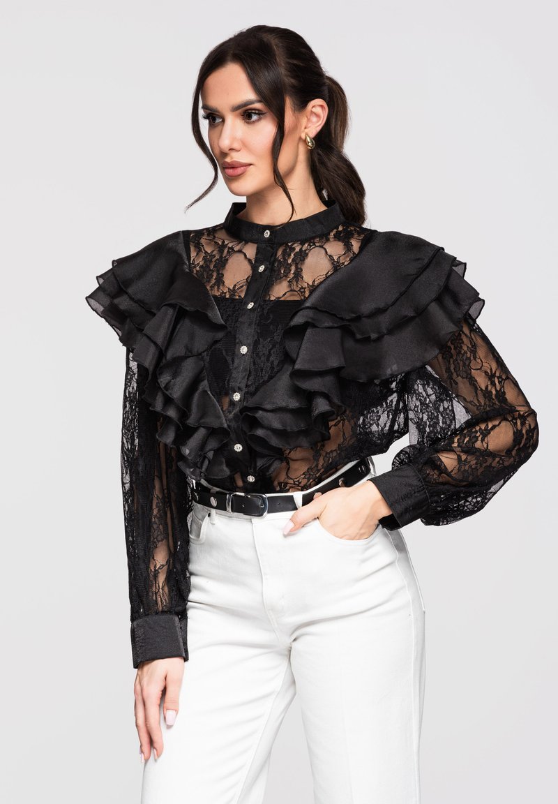 Femme portant un chemisier noir en dentelle transparente à volants superposés, avec des boutons sur le devant, associé à un pantalon blanc taille haute et une ceinture noire, la main dans la poche.