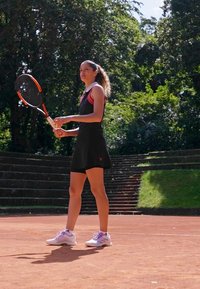 Černé sportovní šaty s růžovými akcenty, bílé tenisové boty a tenisová raketa. Venkovní antukový kurt s zelení a kamennými schody v pozadí.