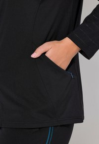 Svart träningstopp med en sidficka med blå dragkedja, slät textur och långa ärmar med vertikala accentstripor.