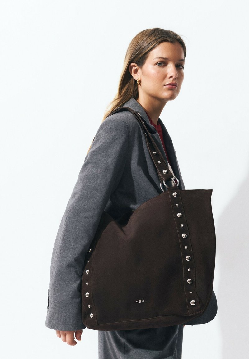 Mujer joven vestida con un traje gris sostiene un gran bolso de hombro marrón oscuro con tachuelas contra un fondo blanco.