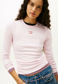 Top de manga larga de raya en color rosa claro con cuello redondo, que presenta un pequeño parche de logo y puños a rayas en contraste, combinado con denim azul.