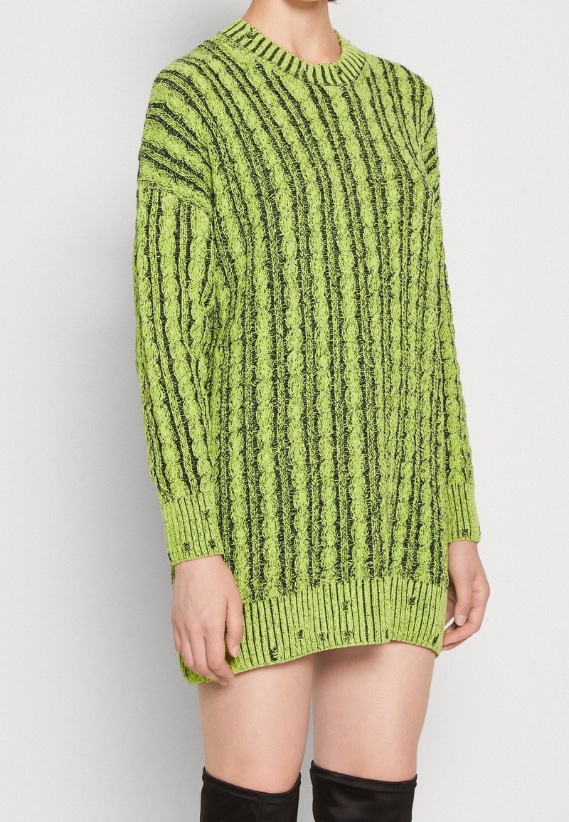 Personne portant une longue robe-pull en maille rayée vert lime et noir à manches longues et des bottes noires hautes jusqu'aux cuisses.