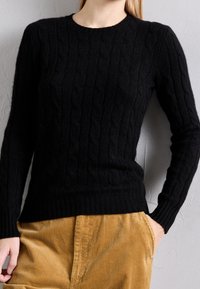 Schwarzer, gestrickter Pullover mit Zopfmuster, rundem Ausschnitt und gerippten Bündchen und Saum. Kombiniert mit hellbraunen Cord-Hosen.