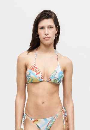 Młoda kobieta w kolorowym bikini z bocznymi wiązaniami z tropikalnymi i tekstowymi wzorami, stojąca na jednolitym jasnoszarym tle.