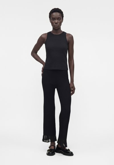 Γυναίκα που στέκεται φορώντας μαύρο αμάνικο τοπ, μαύρο πλισέ παντελόνι με κρόσσια στα τελειώματα και μαύρα μοκασίνια πάνω σε λευκό φόντο.