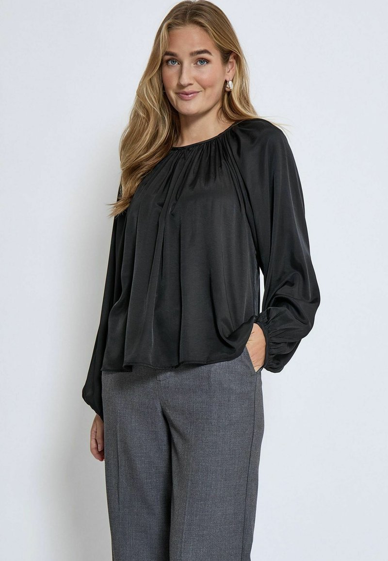 Schwarze Bluse mit langen Ärmeln, plissiertem Vorderteil, runder Ausschnitt und gerafften Manschetten. Kombiniert mit grauen, maßgeschneiderten Hose, die eine lässige Passform zeigt.