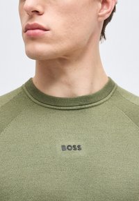 Κοντινό πλάνο ενός ατόμου που φοράει πράσινο πλεκτό πουλόβερ με ριμπ λαιμόκοψη και μαύρο λογότυπο "BOSS" στο στήθος.