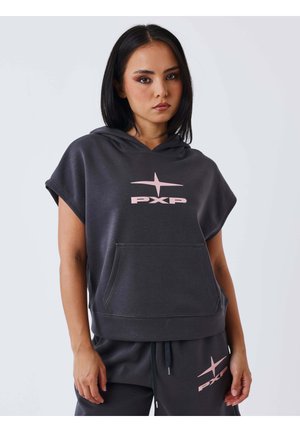 Vrouw met steil donker haar draagt een zwarte mouwloze hoodie en bijpassende broek met roze PXP-logo, staand tegen een effen witte achtergrond.