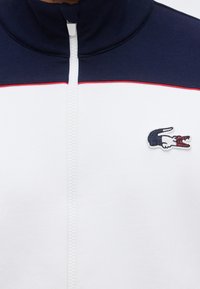 Weiß- und marineblaue Jacke mit Reißverschluss, roter Zierlinie und einem kleinen, gestickten Krokodil-Logo auf der Brust.