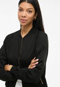 Zwarte bomberjack met een rits aan de voorkant, ribbelkraag en boorden. Stoere stof met een relaxte pasvorm. Gouden ritsdetails versterken het ontwerp.