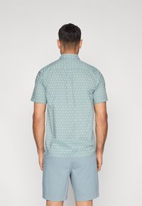 Πουκάμισο με κοντά μανίκια, κουμπιά μπροστά, σε ανοιχτό μπλε χρώμα με μικρές λευκές πουά, με ραφή πίσω και διακριτή υφή. Φορεμένο με ανοιχτό μπλε σορτς.