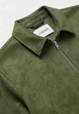 Olivengrønn semsket jakke med krage og sølvfarget frontglidelås, med en hvit etikett med merkenavnet "CISZERE" inne i kragen.