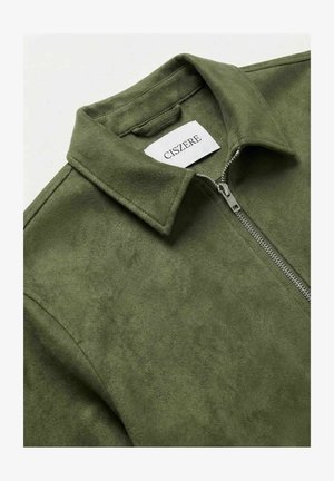 Olivengrønn semsket jakke med krage og sølvfarget frontglidelås, med en hvit etikett med merkenavnet "CISZERE" inne i kragen.