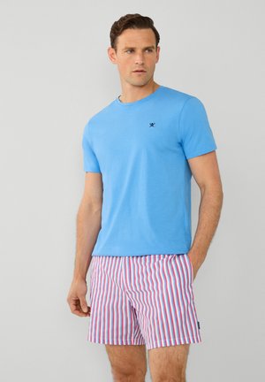 Helles blaues Baumwoll-T-Shirt mit kurzen Ärmeln, versehen mit einem kleinen schwarzen Logo. Kombiniert mit gestreiften Badeshorts in Pink und Blau.