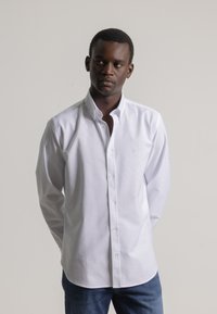 Hombre vestido con una camisa blanca de botones y jeans azules, de pie con las manos detrás de la espalda, mirando de lado contra un fondo liso.