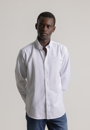 Hombre vestido con una camisa blanca de botones y jeans azules, de pie con las manos detrás de la espalda, mirando de lado contra un fondo liso.