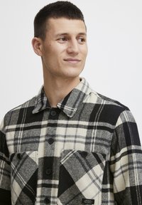 Svart og krem rutete skjorte laget av vevd stoff, med krage, knappelukking og to frontlommer med knappdetaljer.