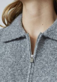 Grå uldjakke med krave og lynlås fortil. Stoffet har en blød, struktureret overflade i en neutral farvetone.