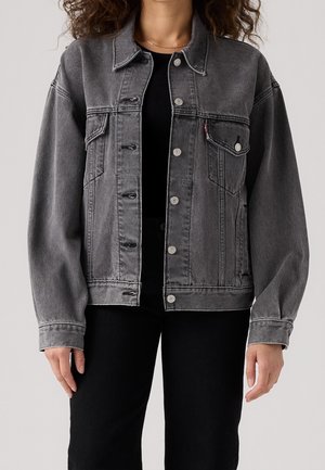 Kvinde iført en falmet sort denimjakke med knapper, brystlommer med klapper og sorte bukser mod en ensfarvet baggrund.