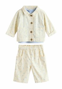 Costum format din două piese, de un galben deschis, ce include o jachetă cu guler, dotată cu nasturi din lemn și pantaloni scurți asortați, ambele decorate cu un mic model floral albastru.