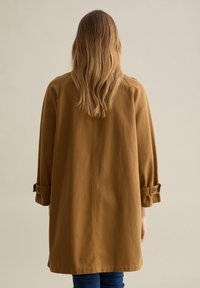 Brauner, mittellanger Mantel aus leichtem Stoff, mit aufgerollten Ärmeln und Knopfakzenten sowie geradem Schnitt.