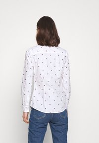 Vit långärmad skjorta med små röda och blå logotyper, böjd fåll, figurnära passform och knappade ärmmuddar, tillsammans med blå jeans.