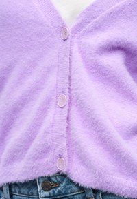 Μαλακό μωβ πλεκτό ζακέτα με κουμπιά στο ίδιο χρώμα, φορεμένη πάνω από ένα λευκό τοπ, συνδυασμένη με μπλε τζιν τζινάκι στη μέση.