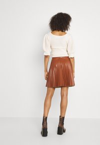 Kvinna med lockigt hår iklädd en vit, texturerad topp, brun veckad kjol och bruna klackade ankelboots som står mot en enfärgad bakgrund.