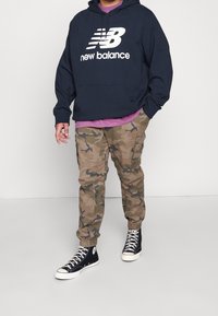 Henkilö, jolla on yllään tummansininen New Balancen huppari, violetti paita, maastokuvioiset cargo-housut ja mustat korkeavartiset tennarit, seisoo vaalealla lattialla.