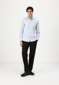 Chemise à manches longues bleu clair avec des boutons sombres, associée à un pantalon noir et des chaussures noires. Le modèle se tient devant un fond neutre.