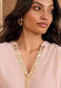 Light pink blouse with a V-neckline and a sequined trim in gold. Smooth fabric with a subtle sheen, featuring a relaxed fit.