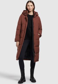 Bruine lange gewatteerde jas met een capuchon, voorzien van een ritssluiting, doorgestikte patronen en zijzakken, gedragen met zwarte legging en enkellaarsjes.