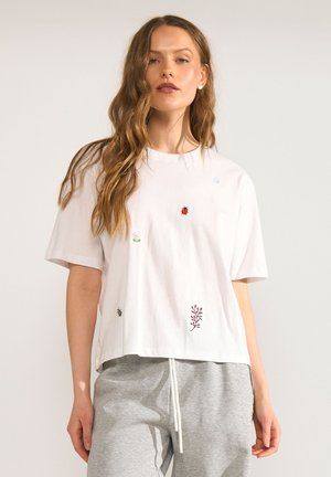 Jeune femme aux longs cheveux ondulés porte un T-shirt blanc avec de petits insectes et plantes brodés ainsi qu'un pantalon de survêtement gris.