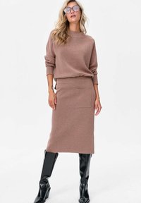 Brązowy sweter dzianinowy z okrągłym dekoltem, w połączeniu z dopasowaną spódnicą midi z boczną kieszenią. Błyszczące czarne kozaki za kolano dopełniają wygląd.