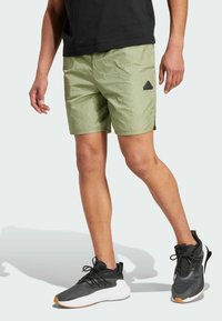 Pantaloni corti verde chiaro con una texture increspata, dotati di uno spacco laterale e un logo adidas nero sul lato sinistro, abbinati a scarpe da ginnastica nere.