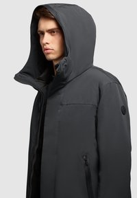Polstret sort parka-jakke med en stor hætte, frontlynlås og sidelommer. Tekstureret stof med et logo-accent på ærmet.