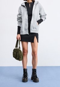 Persona che indossa una felpa grigia con cerniera sopra un vestito nero con spacco sulla coscia, tiene una borsa verde oliva e indossa stivali neri con lacci.