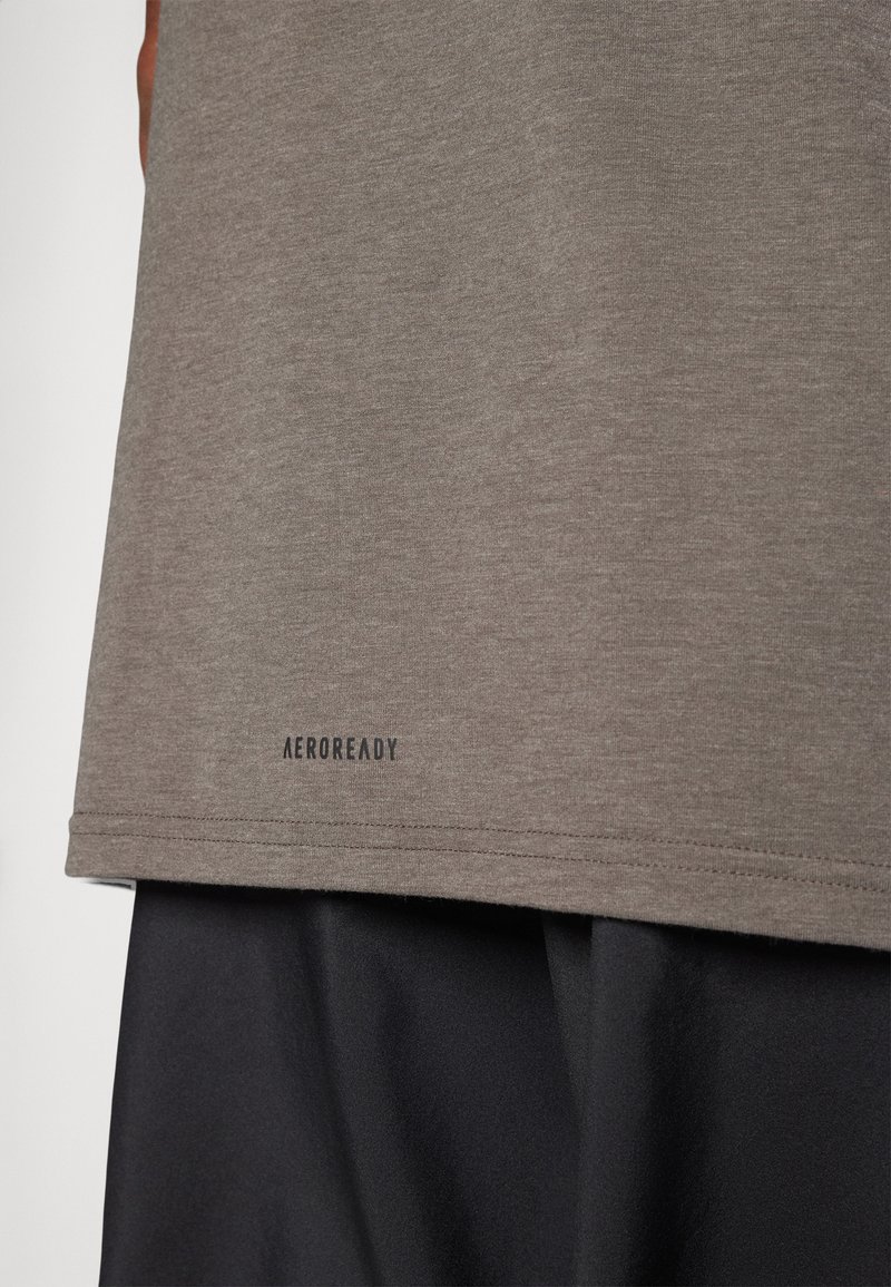 Camiseta deportiva gris con una textura suave, con un dobladillo redondeado y un logo negro de "AEROREADY" en el borde inferior. Combinada con shorts negros.