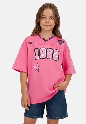 Roze oversized jersey met korte mouwen, met het nummer "1982" en stergrafieken. Het heeft een V-hals en een contrasterende blauwe bies.