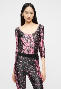 Γυναίκα με μακριά σκουρόχρωμα μαλλιά, φοράει ροζ και μαύρο μπλουζάκι με μοτίβο ψαροκόκκαλου μακρυμάνικο και ταιριαστό παντελόνι με μαύρη ζώνη στη μέση.