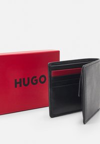 Portofel din piele neagră cu multiple sloturi pentru carduri, aflat parțial deschis, lângă o cutie roșie cu logo-ul negru "HUGO" prominent.