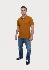 Short-sleeve polo shirt in golden brown with a blue collar accent, paired with blue jeans and navy sneakers, featuring a small logo on the chest.