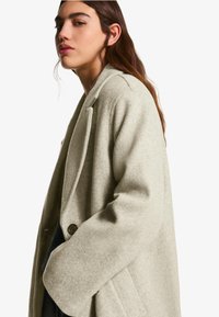 Mladá žena s dlhými hnedými vlasmi, oblečená v svetlošedom vlnenom kabáte, pozerá sa do strany na pozadí jednoduchej bielej steny.