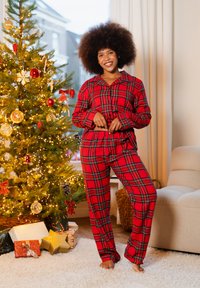 Pyjama en flanelle rouge à carreaux avec une chemise boutonnée et une taille élastique, présenté dans une pièce lumineuse à côté d'un sapin de Noël décoré.
