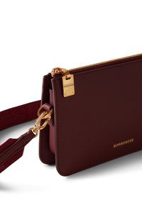 Sac à bandoulière en cuir bordeaux avec une fermeture éclair dorée, des accents matériels dorés et une bandoulière tressée. Présente un logo de la marque embossé d'un côté.