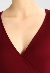 Burgundowa bluzka z zakładkami z głębokim dekoltem w kształcie V, o miękkiej fakturze materiału i starannych detalach szycia. Minimalistyczny design bez widocznych ozdób.